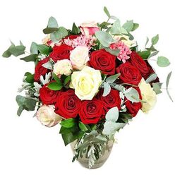A rich, romantic rose arrangement is displayed in a simple clear glass vase, set cleanly against a white studio backdrop so that only the flowers command attention. The bouquet overflows with deep red and vivid scarlet roses, some just beginning to open, others fully unfurled in lush, layered spirals, conjuring the feeling of a passionate declaration offered on a quiet evening in central Oxford. Scattered throughout the crimson sea are luminous white and creamy ivory roses, several with delicate pale pink tips that soften the composition and suggest tenderness and new beginnings. Between the larger blooms, tiny bright pink flowers, likely miniature carnations or similar filler blossoms, add a playful pop of colour and a fine-textured contrast to the smooth rose petals. Flowing through the design are generous lengths of eucalyptus, their round, blue-green leaves creating a cool, contemporary frame and hinting at that fresh, almost herbal fragrance you might notice drifting from a florist near the Covered Market. Additional dark green foliage and a few silvery-toned leaves give the bouquet depth and structure, shaping it into a rounded, generously packed display that looks impressive from any angle. The vase, with its rounded base tapering to a narrower neck, holds the criss-crossed stems neatly, with the line of clear water visible and still. The lighting is bright and even, with no distractions, allowing the colours to glow. This is the kind of arrangement that would sit beautifully on a dining table in North Oxford, grace a wedding anniversary at an Oxford college, or arrive as a memorable same-day delivery from a trusted local florist.