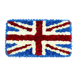 A solemn rectangular floral arrangement, designed as a three-dimensional interpretation of the Union Jack, fills the frame against a simple white backdrop, its colours and details standing out with clarity. The flag is formed entirely from small, closely massed blooms that create a dense, cushion-like surface, transforming a familiar national symbol into a living tapestry. At the centre, a bold red St George's Cross is carefully constructed from vivid red blossoms, their rounded heads nestled tightly together to form smooth, straight arms running vertically and horizontally. Threaded through these, thinner diagonal bands of the same rich red trace the lines of St Patrick's Cross, each edge crisply defined by slender white floral borders. Surrounding this intricate overlay, the broader diagonal white of St Andrew's Cross slices cleanly through the composition, its petals giving off a soft, luminous quality that contrasts beautifully with the surrounding blue. The corners and outer edges are saturated with deep blue flowers, likely compact hydrangea or similar blooms, providing a velvety backdrop that makes the red and white sections appear almost to rise from the surface. Seen as a whole, the tribute radiates both respect and patriotism, suggesting a funeral piece created to honour a loved one's service, roots, or lifelong affection for the United Kingdom. This kind of floral flag is often chosen by families across Oxford, from Jericho to Summertown and down towards Botley, for poignant services in local churches, crematoria, or chapels. The careful balance of colour, the precise geometry of the crosses, and the tactile texture of the clustered flowers all speak to expert local floristry and a desire to create a lasting, dignified gesture of remembrance.