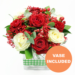 A richly detailed red and white bouquet stands out sharply against a pure white studio background, showcasing every nuance of the florist's work. Full-headed red roses, dark and velvety, are arranged in generous clusters, their petals curling open in elegant spirals. Nestled among them, creamy white roses provide a soft contrast, their ivory tones calming the intensity of the crimson. Between these classic blooms run delicate sprays of tiny red filler flowers that look like tinted baby's breath, adding a frothy, playful texture above the foliage. Glossy red hypericum berries punctuate the arrangement, round and polished, evoking a festive, almost Christmas-like charm that would feel perfectly at home in an Oxford living room overlooking Christ Church Meadow or along the Botley Road. The flowers are supported by a bed of lush greenery: broad dark leaves, sprigs of eucalyptus with a subtle silvery hue, and smaller foliage that rounds out the silhouette. Everything is gathered into a sleek, clear rectangular glass vase, the stems visible through the transparent sides in orderly green lines. A green and white gingham ribbon wraps around the vase's midsection, adding a touch of rustic comfort reminiscent of local farm shops around the Oxfordshire countryside. At the lower right corner of the image, a bold orange circle features the words "VASE INCLUDED" in white, confirming that the display arrives ready to set straight on a table or desk. The bouquet radiates thoughtful celebration, perfect for marking an anniversary, sending love to a student in halls, or brightening a workplace near Oxford city centre.