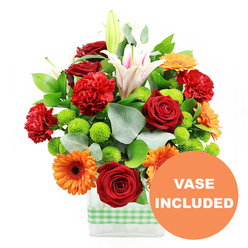 This image shows a joyful, garden-fresh floral arrangement styled in a clear rectangular vase, the sort of bouquet you might see on a sunny windowsill in an Oxford terrace overlooking the River Thames. Around the base of the vase, a light green and white gingham ribbon is tied in a neat band, giving the display a relaxed, cottage-inspired feel that contrasts beautifully with the modern, clean lines of the glass. The bouquet itself is a rich tapestry of colour and texture: velvety deep red roses and plush red-orange carnations create a romantic, full-bodied centre, their petals layered and slightly ruffled. Around them, bright orange gerbera daisies with dark, almost black centres tilt their faces outward, adding a bold, contemporary note that feels as lively as a stroll through Oxford's bustling city centre. White Oriental lilies rise above, some fully unfurled to reveal inner blush-pink stripes and prominent stamens, others still in smooth, tapered buds, suggesting fresh blooms still to come. Scattered throughout, small, round lime green button chrysanthemums form little cushions of colour, bringing a youthful energy that plays against the darker greens of the foliage. Glossy leaves and airy sprigs of silver-green eucalyptus frame the flowers, extending slightly beyond the main mass to give a soft, natural silhouette. The stems are visible through the water, neatly arranged. In the lower right corner sits a bright orange circular badge, overlaid on the image, with the words "VASE INCLUDED" in bold white letters, making it clear that this floral gift arrives complete and ready to enjoy, perfect for same-day delivery to homes, colleges, and offices across Oxford.