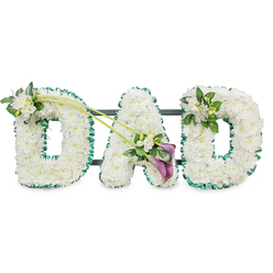 This image shows a carefully crafted funeral flower tribute that spells "DAD" in bold, upright letters, resting against a bright white background so every detail stands out. Each letter is densely filled with immaculate white chrysanthemums, or similar small button flowers, their tightly layered petals forming a plush, almost cushion-like surface that suggests softness and care. Encircling the outside of every letter is a vibrant deep green ribbon, neatly pleated to give a ruffled edge, which adds structure, contrast, and a quiet echo of the greenery found in Oxford's parks and riverside walks. Behind the flowers, a dark supporting frame and horizontal bars can just be made out, indicating that the piece is mounted on a stand, as it might be when placed beside a coffin or at a graveside service. The top left of the first "D" is adorned with a dainty cluster of white freesias and fresh foliage, their slender buds and open blooms resting lightly on the white chrysanthemum base. Two sweeping green stems arc from this first letter across towards the central "A", where they form a graceful diagonal line that draws the eye. At their meeting point sits an expressive spray of flowers: two rich, deep purple calla lilies with sculpted, trumpet-like heads, tucked among more white blossoms and glossy leaves, offering a touch of solemn elegance and colour. On the upper right of the final "D", another cluster of white blooms and greenery mirrors the first, creating a sense of balance. The tribute feels calm, respectful, and deeply affectionate, the kind of personalised farewell a family in Oxford might choose to remember a devoted dad with quiet pride.