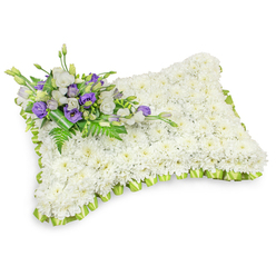 A carefully composed floral pillow, shaped like a comforting rectangular cushion and composed of hundreds of small, immaculate white chrysanthemum heads, creates a serene field of soft texture that invites quiet reflection. The chrysanthemums are arranged so densely that the surface appears seamless and plush, evoking the smooth hush of a resting place. The pillow's margin is defined by a delicately pleated strip of pale lime-green ribbon, which traces the outline with a subtle, hopeful brightness and gives the design a refined, finished edge. Resting across the upper-left corner is an elegant accent spray: crisp white lisianthus with their layered petals mingle with rich purple lisianthus buds, the purple notes offering a restrained, comforting contrast that reads as dignity and remembrance. Intermingled fern fronds and glossy green foliage add depth and a natural breath to the composition, while a neat band of darker green leaves holds the cluster together at its base. Photographed on a pristine white background with gentle studio lighting, soft shadows beneath the pillow hint at its gentle volume and meticulous craftsmanship. The overall mood is one of reverent calm - tactile, understated, and profoundly sympathetic. As a florist familiar with Oxford's funeral customs, I would place a tribute like this at a chapel near the Covered Market or beside a graveside in Headington, where families favour delicate, personal gestures. This design is suited to wakes and memorials and is available for careful local delivery across Oxford and nearby districts.