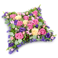 In this detailed image, a plush cushion of flowers appears to float against a bright white backdrop, creating a serene, almost chapel-like atmosphere. The tribute is shaped like a soft rectangular pillow with rounded corners, completely covered in closely set blooms so that no base is visible. Fuchsia and pastel pink roses are the most eye-catching, their petals unfurling in spirals of colour beside creamy lemon-tinged roses that add warmth and light. Nestled between them are clusters of deep purple lisianthus, their velvety trumpets and ruffled edges introducing a gentle, contemplative mood, reminiscent of quiet moments in Oxford's historic churchyards off the High Street or near Jericho. Delicate sprays of tiny lavender-hued flowers, likely statice or limonium, weave through the design, lending a misty, cloud-like softness. Around and beneath the blooms, cool, rounded eucalyptus leaves in silvery green catch the light, while darker, finer foliage adds shadow, dimension, and a sense of natural woodland texture. The flowers are arranged so densely that the surface looks cushioned and comforting, as though it could cradle a final message of love. The lighting is soft and even, emphasizing the intricate textures of each petal and leaf without harsh shadows. This floral pillow is a classic sympathy design, often chosen in Oxford for funeral services, cremations, and graveside tributes, where families wish to convey deep affection, gratitude, and a peaceful farewell through a tangible, beautifully crafted arrangement of fresh, seasonal flowers.