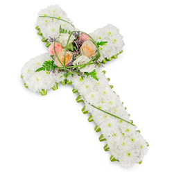 A traditional Christian cross crafted entirely from flowers rests against a clear white background, its quiet elegance suited to funerals and memorials across Oxford. The structure of the cross is generously padded with tightly packed white cushion chrysanthemums or similar small blossoms, arranged so precisely that they form a smooth, velvety surface reminiscent of fresh snow along the paths of Port Meadow. Each petal contributes to a sense of stillness and purity, inviting reflection and gentle remembrance. Around the perimeter of the cross runs a narrow, vivid green edging of foliage or small green floral pieces, hugging every curve and corner so the shape remains crisp and defined against the pale backdrop. At the centre where the arms meet, a raised floral spray introduces soft colour and emotional warmth. Several roses in full bloom, in tones of soft peach, pale blush pink, and creamy white, are clustered together like a tender embrace. Their rounded forms contrast with the tiny white blooms beneath, creating a natural focal point. Wispy green ferns, fine strands of looping grass, and discreet accents of tiny purple filler flowers weave among the roses, adding movement and texture without overwhelming the design. The overall lighting is clean and bright, without glare, allowing the subtleties of cream, green, and lavender to show clearly. This floral cross feels especially fitting for services in local churches near the High Street or quiet chapels in Cowley, a respectful tribute that speaks of love, farewell, and the hope of peace beyond this life.
