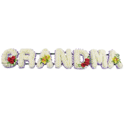 This image shows a lovingly constructed GRANDMA funeral tribute, laid out in bold capital letters against a bright white background so the flowers appear luminous and clear. Each letter is completely covered in tight clusters of white chrysanthemums, their many petals overlapping to create a soft, cushion-like feel, as if the word itself has been wrapped in a gentle embrace. Just beneath the white blooms runs a narrow band of light lavender flowers, adding a misty violet outline that gives depth and definition to the shapes without disturbing the peaceful mood. Subtle accents of colour bring the piece to life. The letter G carries a cluster of three deep red roses at its lower curve, their rich tone symbolising love and devotion, framed by dark, polished greenery. The first A features two elegant lime-green orchids with warm reddish markings at their centres, resting across the bar of the letter and introducing an exotic note. The letters R, N, and D remain purely white with their lavender edge, calm and undistracted, while the M holds another trio of red roses tucked into its lower right side. On the final A, two more yellow-green orchids mirror those on the first, balancing the design from end to end. A slim dark green or black metal frame links the letters securely behind the blooms, ready to rest on a stand beside a coffin or at the front of a chapel. The arrangement feels composed and respectful, the sort of bespoke tribute a family around Oxford's quiet neighbourhood churches or cemeteries might select to remember a gentle, much-missed grandmother.