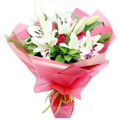A luxurious bouquet of white Oriental lilies and red roses is captured in full detail against a plain white backdrop, allowing every nuance of the flowers to shine. At the centre, tall stems of lilies rise elegantly, some flowers fully open with smooth, white petals stretching outward, others still wrapped in elongated buds that blush faintly green at the tips. Their prominent stamens carry dark mahogany to almost purple-black anthers, which rest like tiny brushstrokes of colour inside each bloom. Threaded among these lilies are rich red roses, many still in tight, compact buds that promise to spiral open, giving the design an air of classic romance. Around the blooms, a generous collar of dark and mid-green foliage creates a dense, natural frame; glossy broad leaves catch soft reflections, while slender, spiky greenery adds texture, almost echoing the wild hedgerows that line Oxford's side roads near Cowley and Headington. The bouquet is wrapped in a billowing layer of bright pink cellophane or floral film, shiny and semi-transparent, gathered into pleats and folds that glisten in the light like delicate silk. At the base, a slim strip of lime green ribbon is tied snugly around the stems, a fresh, zesty accent that unites the vivid wrapper with the deep green leaves. The overall effect is joyful yet refined, an arrangement that could easily be carried down a cobbled street off Cornmarket for a birthday surprise, or delivered across Oxford as an elegant expression of love, gratitude, or celebration.