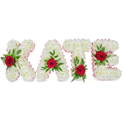 An intimate floral tribute spells out KATE in large sculpted block letters, each letter lovingly built from dense clusters of small white chrysanthemums that give a soft, cushiony texture and a sense of quiet dignity. The edges of the letters are traced with a subtle pale pink satin ribbon that catches light and introduces a whisper of colour, framing the name with tenderness. On the face of every letter a single vivid red rose is deliberately placed amid sprigs of lush green foliage - broad leaves and feathery ferns that create depth and cradle each bloom. The placement is carefully considered: the rose on the K settles low on its left vertical stem, the A is crowned with its rose at the triangular apex, the T is anchored by a crimson bloom at the base of the left side of its crossbar, and the E reveals its rose along the middle horizontal arm. The arrangement rests on a clean white background, the white chrysanthemums symbolising purity and remembrance while the red roses speak of deep affection; together they compose a composition both classic and personal. As an Oxford florist might imagine it being received, the piece feels appropriate for a chapel near the Radcliffe Camera, a funeral at the university church, or a family memorial service - a handcrafted, emotive statement designed to console and honour a life remembered.