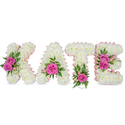 A carefully crafted name tribute spells KATE in freestanding, three-dimensional letters, each sculpted from hundreds of small, immaculate white chrysanthemums that form a dense, cloud-like texture. The white letters are neatly edged inside with a pale pink satin ribbon that catches the light and subtly defines each letter's inner shape; soft shadows on the white backdrop enhance the three-dimensional quality. Bright hot pink roses are used as intentional focal points-two fully opened roses accent the left side of the K, a single bold rose anchors the lower curve of the A, the T is graced with one vivid rose at its base, and the E shows a cluster of two pink roses along its right-hand strokes. Small pale-pink buds, possibly alstroemeria sprays, and a mix of fresh green foliage-variegated leaves, fern-like fronds, eucalyptus and ruscus-are woven around the roses to add texture, colour contrast and a subtle herbaceous scent. The chrysanthemums are packed close for a seamless surface, while the ribbon and foliage give structure and a delicate finish. As florists in Oxford know, this kind of bespoke name tribute reads clearly at services in venues from the Covered Market chapels to University Parks memorial events; Flowers Oxford supplies similar personalised tributes with care and local delivery across Jericho, Headington and Cowley. The arrangement's tone is reverent and loving, a dignified visual message of remembrance and celebration of a well-loved life.