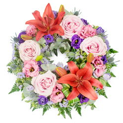 Centred on a crisp white background, this circular sympathy wreath appears almost like a painted halo of flowers, rich with detail and feeling. Imagine it resting with quiet dignity in a chapel near Oxford's Jericho or at a service in Summertown: the composition is full, expertly constructed, and deeply respectful. Two bold Asiatic lilies in a warm reddish-orange hue anchor the design, one placed toward the top of the ring and the other near the bottom, their wide petals flaring open to show dark stamens. Their colour brings a vivid, sunset-like glow against the softer pastels, symbolising strength and love that remain even in times of loss.   

Surrounding these lilies are generous clusters of open roses in blush and soft pink-lavender tones, petals layered in tight spirals that feel almost cloud-like. Threaded through them are lisianthus blooms, some in pale pinks and others in velvety deep purples, their gently ruffled edges introducing a romantic, almost whispering texture. Among the larger flowers, small sprays of lavender-blue filler flowers, similar to statice or limonium, form delicate clouds of colour, softening transitions and guiding the eye around the wreath. Spiky blue eryngium thistles add cool, silvery-blue accents, their sculptural forms lending quiet resilience to the tribute.   

Beneath the blossoms, a lush framework of foliage creates a sense of abundance: silvery-green eucalyptus, airy fern fronds, and deeper green leaves weave together seamlessly. Bright green, folded leaves near the focal lilies give the design a modern, tailored touch, the kind an experienced Oxford florist would shape for a bespoke funeral arrangement. A few closed lily buds nestle among the greens, promising future blossoms and suggesting that love and memory continue beyond the day of the service.