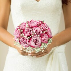 This evocative close-up captures the quintessential spirit of an Oxford wedding, with a bride's poised hands gently enveloping a radiant bouquet that practically sings with local artistry. At the heart are roughly a dozen pink roses, each masterfully arranged so their petals blossom outwards in a brilliant ombré, their inner blush soft as morning light over Christ Church Meadows, fading to sumptuous fuchsia and magenta at the edges. Tiny, velvety white baby's breath weaves between the roses, adding a dewy freshness and a lace-like delicacy that echoes both the detailed bridal gown and the historic craftsmanship found along Oxford's High Street. The bride's sleeveless white dress, its bodice richly textured in classic lace with a modern V-neckline, is complemented by a softly tied sash off to the right-its subtle drape a gentle nod to the city's timeless elegance. Neatly manicured nails, painted in a pale, understated sheen, add refinement to the natural warmth of her tanned skin. The entire composition, suffused in neutral, gentle light, directs full attention to the bouquet-a floral work of art that embodies the dreams and excitement of a wedding day in Oxford. Such blooms, carefully chosen for their colour and scent, might easily be handpicked from the bustling stalls of the Covered Market or sourced fresh from Flowers Oxford, ensuring an unforgettable start to a lifelong journey.