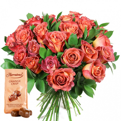 As an Oxford florist deeply attuned to the locality's unique rhythm, I see this spectacular gift ensemble as a symphony of colour and connection. The rounded bouquet takes centre stage, bursting with roses in an array of luminous shades-from the gentle glow of peach and buttery yellow at each petal's heart to robust, sunset-inspired hues of salmon, coral, and nuanced pink that recall the changing light across Port Meadow. The foliage, fresh and dark green, deftly frames the roses like the elegant gardens bordering Christ Church. Each bloom's texture is palpable, from the lush softness of their open petals to the glossy resilience of the surrounding greenery-a feast for both eyes and fingers. Completing the sensory journey, a Thorntons Orange Crisp chocolate pouch anchors the bouquet, its creamy beige wrapping and inviting typography familiar from Oxford's favorite high street confectioner. The packet, illustrated with two glossy chocolate truffles, promises a tangy, zesty filling and the crunch of crisp wafer, deliciously finished in milk chocolate. Two unwrapped truffles rest before the pack, rich and decadent, ready to be savoured. Whether celebrating an academic milestone, rekindling romance along Cowley Road, or sending comfort across leafy Summertown, this bouquet and chocolate pairing encapsulates the warmth, luxury, and generosity that make Oxford's gifting traditions so cherished.