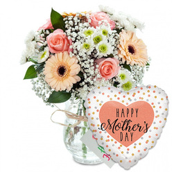 This Mother's Day ensemble is captured against a spotless white backdrop, where every blossom and reflective surface feels calm and intentional, like a still moment before family gathers for lunch in Summertown. The focal point is a lush, rounded bouquet resting in a clear glass vase, its sides smooth and simple, tied at the neck with a casual twist of pale twine that hints at countryside charm beyond Oxford's city streets. Within the bouquet, several peach-pink roses are fully open, their layered petals forming delicate spirals that invite you to lean in, while two or three generous gerbera daisies in warm peach-orange tones, each with a velvety dark brown centre, add a bold, sunlit presence. Between these stars of the arrangement, clusters of white baby's breath float like tiny clouds, and compact white chrysanthemums with subtle green hearts bring both softness and structure. Rich green foliage curves around the flowers, framing them as if they were gathered from a well-tended garden off Cowley Road. Nestled behind the blooms, almost hidden, sits a small white greeting card with a splash of green and pink handwriting, suggesting a heartfelt Mother's Day message from a child or partner. To the right of the vase, a large heart-shaped foil balloon leans forward with playful grace. The balloon's bright white surface is sprinkled with metallic gold and silver dots, mimicking scattered confetti, while a central peach heart carries the message "HAPPY Mother's DAY" in bold black letters, the word "Mother's" highlighted in flowing, calligraphic script. The combination of fresh flowers, thoughtful card, and celebratory balloon creates an image of warmth, gratitude, and gentle festivity, ideal for a same-day Mother's Day delivery across Oxford's neighbourhoods.