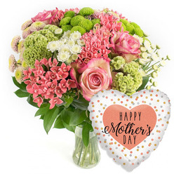 A lavish Mother's Day arrangement glows against a simple white backdrop, much like a bright shop window on Cornmarket Street catching the afternoon light. The flowers are gathered into a rounded bouquet and presented in a clear glass vase, the clean lines of the glass allowing the stems and clear water to show, hinting at the care taken by an experienced Oxford florist. At the heart of the design, several large pink roses unfurl, each petal gently curling and edged with a slightly darker hue, creating a soft, romantic focal point. Around them cluster small coral-pink flowers reminiscent of kalanchoe, tightly grouped to create pockets of texture and warmth. Lime-green button chrysanthemums stand out with their compact, neatly domed heads and fine, feathery petals, bringing a bright, modern twist to the otherwise classic palette. Interwoven through the bouquet are airy white spray chrysanthemums, their daisy-like faces adding lightness and a sense of space, while tiny spiky coral blooms and occasional chartreuse green florets tuck into the greenery, adding layers of interest when viewed up close. Rich, dark green foliage cradles the blossoms, framing the colours and giving the impression of a carefully curated garden in miniature, the kind you might discover in a quiet courtyard off St Giles'. To the lower right, a heart-shaped foil balloon completes the gift. Its glossy surface reflects the light, and its white or pale silver ground is dotted with shimmering gold, silver, and peach confetti-style spots. In the centre, a peach heart bears the message "HAPPY Mother's DAY" in black script and bold type. The overall mood is one of festive gratitude and affection, a ready-to-deliver Mother's Day surprise for homes across Oxford, from Summertown to Cowley.