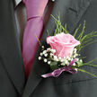 This image captures a refined moment of preparation, focusing closely on the chest and shoulders of a person dressed for a special occasion in Oxford, perhaps on their way to a ceremony near the Bodleian or a reception tucked away off the High Street. The dark charcoal or black suit jacket is sharply cut, its left lapel providing a stage for a thoughtfully crafted boutonniere. Beneath the jacket edge, a pale dress shirt with a soft, checked pattern peeks through, its light texture contrasting gently with the gleaming fuchsia tie that cascades down the centre of the frame. The tie's satin sheen catches the light, its rich magenta tone providing a modern, spirited splash of colour. Pinned carefully to the lapel, the floral design draws the eye: a single, perfectly opened light pink rose, petals rounded and velvety, shading from a deeper blush at the base to tender, almost pearly pink at the tips. Surrounding this central rose, clusters of tiny white baby's breath create a halo of softness, each minuscule flower adding to an overall impression of delicacy and romance. Slender arcs of bright green foliage rise from behind, like fine grasses or herbs you might see in a local Oxfordshire garden, giving the boutonniere height and a gentle, organic energy. At the base, the stems are neatly wrapped in a darker green leaf or ribbon, secured with a slim, satiny lavender bow that mirrors both the rose and tie without overpowering them. The result is a boutonniere that feels personal and expressive, the kind of piece Flowers Oxford might create for couples marrying in city churches or historic college chapels, where every petal and ribbon is chosen to honour the promise of a new beginning.