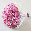A beautifully composed bouquet of roses lies diagonally across a pale, softly creased fabric, the neutral beige background allowing every floral detail to stand out, much as it might on a linen-covered table at an intimate Oxford reception. The arrangement is centred on a cluster of plush roses, their petals moving from an inner haze of lavender-pink and dusty mauve to bold fuchsia and deep magenta tones at the outer edges. Each rose appears either closely cupped or just opening, revealing a careful swirl of petals that suggests both structure and tenderness. Nestled closely around and between these pink blooms is a full collar of white baby's breath, the fine Gypsophila blossoms forming a misty, star-like border that enhances the sense of romance. A few dark, glossy green leaves punctuate the softness, adding a natural, garden-fresh dimension. Toward the bottom right of the image, the exposed stems are gathered together and wrapped in a smooth lilac satin ribbon, which gleams gently under the light. Twined around this ribbon is a delicate string of tiny white pearls, evoking the refinement of a bridal bouquet chosen for a ceremony in one of Oxford's historic chapels or a celebration overlooking the dreaming spires. The lighting is gentle and even, picking up the subtle purples, pinks, and creamy whites, while the texture of the fabric beneath echoes the bouquet's softness. The overall mood is serene and romantic, perfect for marking engagements, weddings, or anniversaries, and it feels like a design a local Oxford florist would create for a couple who love understated elegance.