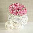A dome of pink roses, lush and full, rises from a sculptural white orb vase in this elegant wedding composition, evoking the calm of an early morning stroll through Oxford's University Parks. Each rose is carefully placed so its petals open in soft spirals, transitioning from a gentle blush at the outer edge to a richer magenta in the centre, as though brushed with watercolour. Around the roses, clouds of baby's breath form a delicate border, their tiny white blooms adding lightness and a hint of vintage charm without disturbing the clean, modern silhouette. The container itself is an open, hand-woven white globe, its criss-crossing strands creating a geometric cage that feels both playful and refined, similar to the contemporary art pieces on display near the Ashmolean. Light plays through its gaps, throwing looping shadows across the tabletop and subtle reflections on the smooth surface below. The setting is bright yet tranquil, with neutral tones that allow the pink and white flowers to take centre stage, much like a carefully curated wedding breakfast table in an Oxford college hall or a boutique venue off Cowley Road. As a florist working regularly on local weddings, I see this design as a centrepiece that speaks of tenderness, poise, and modern romance-ideal for top tables, guest arrangements, or a statement accent beside a signing table, bringing a sense of serene celebration to any ceremony or reception across Oxford.