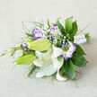 On a soft, off-white linen surface, a horizontally arranged bouquet lies like a small, cultivated patch of meadow. The dominant white Oriental lilies, some fully open with their broad, curling petals and bright orange-yellow stamens, show tiny dark pollen flecks and delicate maroon speckles toward their throats. Between these luminous lilies, white lisianthus edged in violet and deeper purple provide a painterly counterpoint-each ruffled petal moving from a creamy centre to a wash of lavender at the margins. Several lisianthus buds remain tightly furled, promising later blooms and a sense of time within the bouquet's composition. Tiny clusters of Gypsophila (baby's breath) dot the arrangement, lending an ethereal, cloud-like texture that lifts the heavier blooms.
The foliage is varied and deliberately textural: glossy, broad leaves (like salal or ruscus) anchor the shape; slender, feathery foliage and spike-like grasses give direction and movement. The stems are neatly wrapped in a white satin ribbon, completed with small, gem-like pin details that twinkle gently in soft light. The tactile contrasts-silky lily petals, ruffled lisianthus edges, airy gypsophila and crisp greenery-create a bouquet that feels fresh and intentional. It suggests a quiet Oxford morning, as if gathered from a market stall off the High Street and arranged with considered care, perfect for weddings, anniversaries or a thoughtful, elegant gift.