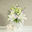 In this thoughtfully composed image, a wedding-ready bouquet of white lilies and lisianthus is placed at the centre of a clean, light-filled setting, evoking the tranquil courtyards and stone cloisters of Oxford colleges. The lilies are the first thing you notice: three or more large, open blooms with velvety white petals that sweep outward, revealing intricate speckles and strong, yellow-orange stamens that suggest a soft, powdery fragrance. Nestled among them, lisianthus flowers in creamy white unfold in layers of gently ruffled petals, some tinged with a delicate lavender outline like a faint watercolour wash, giving the arrangement a subtle romantic glow. Unopened lily buds in pale green rise above the open blooms, adding height and a beautiful sense of expectation, as though the bouquet will continue to evolve over the days ahead. Wispy green foliage and tiny white filler flowers, similar to baby's breath, weave in and around the larger blooms, softening the structure while small clusters of deep blue or purple blossoms provide just enough contrast to make the whites appear even more luminous. The bouquet rests in a distinctive, round vase crafted from white woven strands, creating an airy, basket-like pattern that feels both modern and handcrafted, the sort of piece you might discover in a stylish Jericho home. The vase stands on a pristine white surface, accompanied by two light rattan-style spheres that echo the woven texture and add a natural, earthy detail. A muted beige, finely textured background fades quietly into the distance, letting the flowers take centre stage. The overall impression is one of calm sophistication, ideal for wedding receptions in Oxford barns, elegant hotel ceremonies, or peaceful condolence flowers delivered to families across the city.