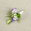 A boutonniere like this speaks the language of quiet romance: two snow-white lisianthus edged in lavender-violet form a tender focal point, their silk-soft petals folded with the deliberate care of a hand florist. Around them, feathery greens and slender fern fronds create a soft halo, while tiny sprays of baby's breath settle into the hollows, lending a whisper of clouded light to the composition. Narrow stems of grape hyacinth run beneath the lisianthus in concentrated ribbons of blue-violet, the compact, bell-like blooms providing a vivid contrast and a slightly peppered scent beneath the sweeter lisianthus aroma. Pale, unopened buds and thin greenery extend with a natural, springlike rhythm, and a neat satin ribbon-tied in a small, reassuring knot-gathers the stems so the piece sits secure on a lapel. Placed against a neutral linen backdrop, the colours read with calm clarity; the woven fabric deepens the tactile story, reminding one of market mornings at the Covered Market or strolls through Jericho when the air feels particularly green. Prepared with the steady hand and attention to detail you'd expect from a local maker, this boutonniere is the kind of delicate accent chosen for vows, portraits, or milestone occasions. Flowers Oxford arranges these small pieces with an eye to balance and longevity, making sure the textures and scent carry the memory of the day long after the jacket is folded away.