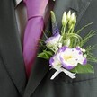 A close-up photograph captures the upper torso of a man dressed for a wedding in Oxford, with the focus resting on a meticulously crafted boutonniere and coordinated formalwear. The suit jacket is a deep charcoal grey, sharply tailored with a clean, structured lapel that frames the floral detail. Beneath it sits a light grey checkered dress shirt, its fine pattern adding quiet texture, while a lustrous magenta silk tie, knotted with care, delivers a bold, confident flash of colour. Pinned to the left lapel is a striking boutonniere built around a white lisianthus bloom edged in rich purple, its ruffled petals opening like a tiny watercolour painting. A second lisianthus and several slender, unopened white buds cluster close, suggesting new beginnings and the gentle promise of the day ahead. Rising behind them, a slim spire of deep indigo-purple veronica draws the eye upward, giving the design an elegant vertical line. Wisps of feathery greenery and small sprigs of baby's breath weave through the flowers, softened by glossy emerald leaves and delicate loops of fine green grass or wire that add a modern, sculpted feel. At the base, a neat white satin ribbon bow gathers the stems into a compact, polished finish. The lighting is soft, almost like natural daylight filtering through an Oxford townhouse window, lending a warm glow to the tie and petals. The overall impression is one of refined celebration, perfect for a groom or groomsman heading to a ceremony by the dreaming spires, where every detail has been chosen to honour love, commitment, and the quiet elegance of the occasion.