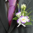 Against the deep charcoal weave of a formal suit, this boutonnière balances classic elegance with a fresh, contemporary edge. The hero is a white lisianthus with a vivid purple rim, its petals layered and softly ruffled so that each edge throws a subtle shadow and catch of light; the effect is both tender and dramatic. Complementing it, a compact white dianthus edged in purple adds textural contrast - its fringed petals bring a spirited rhythm beside the lisianthus's smooth curves. Behind them, three pale-green lisianthus buds stand like hopeful notes, while feathery ornamental grass introduces airy motion and a natural silhouette. A slim stem of deep indigo veronica reaches upward, lending vertical refinement and a cool tonal anchor, and tiny sprays of gypsophila sprinkle bright, cloud-like accents through the arrangement. Dark green foliage cradles the blooms, offering depth and an earthy sheen, and a crisp white ribbon secures everything with understated restraint. Placed on the lapel next to a magenta tie, the piece reads as thoughtfully tailored and quietly joyful - a refined flourish for a groom or an honoured guest. As an Oxford florist I imagine delivering this to a young couple exchanging vows near the Botanic Garden or to a party gathering beneath Carfax Tower; the scent is fresh and floral, never overpowering, designed to reassure through a long, meaningful day. Flowers Oxford prepares these with local expertise and care, available for same-day delivery across central Oxford when timing matters.