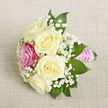 A beautifully composed petite bouquet rests at a diagonal angle on a neutral, light beige textile that looks like finely woven linen, its subtle texture providing a calm, understated backdrop. The focal point is a cluster of five ivory-white roses, each bloom generously petalled and gently opening, their rounded forms giving a sense of softness and quiet luxury. Just left of centre, a single bright fuchsia rose stands out with confident colour, its lively hue immediately drawing the eye and introducing a playful yet romantic accent to the otherwise classic palette. Interlaced throughout the roses are airy sprays of white baby's breath, their countless tiny blossoms creating a misty, cloud-like effect that softens the transitions between the flowers and adds a sense of lightness. Surrounding the arrangement, rich green pointed leaves offer contrast and definition, their deeper tones grounding the composition and hinting at garden freshness. The stems are expertly bound into a compact handle, wrapped in a smooth, lustrous pink satin ribbon that glows softly against the neutral background. Over this ribbon, narrow bands of small, iridescent pearls are arranged in neat horizontal rows, catching the light like tiny dewdrops and elevating the bouquet into something distinctly ceremonial. Captured from a slightly elevated, close perspective with even, flattering light, the bouquet feels intimate and personal-ideal for a bride's maid-of-honour, a prom date walking through Oxford's historic streets, or as a delicate table accent for a celebration in Jericho or near the Botanic Garden, created with the thoughtful precision of an experienced local florist.