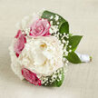 Displayed against a light beige, linen-like fabric that evokes the charm of an Oxford townhouse interior, this bouquet captures the gentle glow of a spring or early summer morning. The central focus is a sumptuous creamy peony, fully open and plush, its layered petals overlapping like pages in a cherished book. Around it, fuchsia-pink roses nestle in close formation, their tight spirals and velvety texture adding depth and a classic romantic note. Threaded among the main blooms are airy clusters of white gypsophila, each tiny blossom forming a star-like accent that diffuses the stronger colours and creates a halo effect around the peony and roses. Rich, dark green leaves arc around the bouquet, some curling protectively at the edges, others lying flatter to frame the design, giving the flowers a sense of structure while still appearing natural and relaxed. The stems are carefully aligned and bound with a pure white satin ribbon, wrapped several times to form a smooth, polished handle that suggests ease and comfort for a bride walking through Oxford's cobbled streets near the Radcliffe Camera or crossing a college quad. The lighting is soft and even, bringing out subtle colour variations in the petals-from creamy ivory to deeper blush tones-and revealing the gentle sheen on the foliage. This arrangement feels intimate and poised, ideal for an Oxford wedding ceremony, a civil partnership at the Town Hall, or a romantic vow renewal, where a refined yet heartfelt style is desired. It radiates quiet confidence, freshness, and the enduring charm of flowers chosen at just the right moment of bloom.