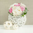 An elegant wedding-style bouquet is showcased on a crisp white surface, its rounded, carefully balanced shape giving it the look of a bridal posy ready to be carried through an Oxford college quad. At the centre, lush ivory peonies open generously, their layered petals forming soft, pillowy rosettes that seem almost weightless, like gathered silk. Around them, rich fuchsia and soft pink roses cluster in tight whorls, each flower at a slightly different stage of bloom, creating gentle movement and depth. The interplay of creamy white and vivid pink draws the eye, while fine sprays of baby's breath thread between the larger blossoms, their tiny white flowers catching the light and introducing a gauzy, romantic softness. Encircling the base, smooth, deep green leaves arc outwards, adding structure, contrast, and an impression of fresh garden foliage snipped that morning from a well-tended Oxfordshire border. The bouquet is cradled in a striking white vase crafted from slender, interwoven strands, like a modern wicker lattice. This open, basket-like construction reveals glimpses of the stems inside and casts subtle shadows on the tabletop, reinforcing a sense of lightness and air. In front of the vase, two decorative orbs made from pale woven material rest casually on the surface, echoing the texture of the vase and giving the arrangement a cohesive, designer feel. The background is a soft, neutral beige, hinting at a linen wall or fabric panel, which keeps the scene calm and understated so the flowers can shine. It feels like a thoughtful centerpiece you might find at a small Oxford wedding reception along the Banbury Road, or a romantic gift delivered to a townhouse near the Covered Market.