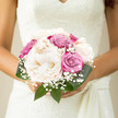 This bridal bouquet feels like a calm pause in the middle of a joyful wedding morning in Oxford, perhaps just before leaving a townhouse near Jericho for the ceremony. Plump, creamy peonies form the glowing centre of the design, their soft ivory petals layered so abundantly that they resemble little cushions of cloud. Nestled closely beside them are roses in dusty pink and gentle violet, their tightly wound hearts beginning to loosen, suggesting a love story that is gracefully unfolding. Wisps of gypsophila, or baby's breath, weave through the flowers in tiny, airy clusters, evoking a light sprinkling of confetti or a trace of morning mist drifting over the Cherwell. Rich green foliage cups the flowers, the leaves firm and smooth, creating a cocoon that highlights the paler tones while adding depth and structure. The bouquet is bound in a way that feels intimate and hand-crafted, sized perfectly to be cradled against a lace gown or simple satin dress. In soft, natural daylight, the whole arrangement seems to glow, radiating clarity, balance, and a sense of serene happiness. You can imagine the faint mix of floral scents-peonies, roses, and fresh greenery-following the bride through the stone archways and cloisters of central Oxford. Ideal for an elegant city wedding, this bouquet captures the harmony between tradition and contemporary romance that couples so often look for when choosing their flowers here.