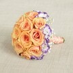 Set against a subtly textured beige backdrop evocative of Oxford's storied libraries, this vibrant round bouquet is a vision of celebration and elegance. The design is dominated by an abundant cluster of peach, apricot, and soft orange roses-each bloom tightly nestled, their petals unfolding with a gentle allure that conjures the warmth of spring afternoons along High Street. Along one graceful sweep, white lisianthus edged in deep violet bring an element of surprise and sophisticated contrast, their ruffled shapes echoing wildflowers spotted in the Botanic Garden. Wisps of pure white baby's breath are artfully scattered throughout the bouquet, infusing the arrangement with buoyancy and an almost ethereal delicacy. The stems are gathered and carefully wrapped in a luxurious, peach satin ribbon, culminating in a polished handle studded with a continuous string of small, creamy pearls-the finishing flourish that speaks of timeless romance and thoughtful, local craftsmanship. Apt for a wedding beneath Oxford's dreaming spires, a milestone anniversary, or a springtime gift delivered to Summertown, this bouquet carries with it the ambiance of refined celebration unique to the city. Evoking both joy and serenity, it represents the ideal blend of tradition and innovation that Oxford's community treasures.