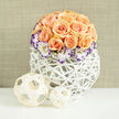 A compact, dome-shaped gathering of apricot and peach roses commands the view, each bloom arranged with the careful touch of an experienced hand so that two to three dozen flowers form a plush, pillowy mass of colour. Some roses open wide, their velvety layers fanning outward, while others hold close in neat buds, creating depth and movement across the bouquet's surface. Nestled around the lower curve of the rose dome, white Lisianthus with cream interiors and richly violet-edged petals introduce a refined, painterly contrast that draws the eye. Wisps of baby's breath are threaded through the arrangement, giving the composition lightness and a breathy, classic romance. The flowers sit in a contemporary rounded white woven vessel - an airy, open-lattice design that resembles bright wicker or reed - which supports and softens the bouquet with its handcrafted texture. Two pale wooden or bamboo decorative spheres rest to the left on the tabletop, their openwork weaving mirroring the vase and adding an organic, sculptural accent. A soft, neutral linen-coloured backdrop blurs into gentle warmth, as if viewed through early afternoon light near Jericho or the University Parks in Oxford. The scent seems almost present: a subtle, fresh rose perfume with a hint of meadow-like sweetness from the Lisianthus and Gypsophila. This arrangement reads as a carefully considered gift - for an anniversary, a cosy housewarming by the High Street, or a thoughtful celebration - where modern design meets gentle, floral sentiment in a way that invites touch and quiet admiration.