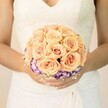 A close-up, chest-level photograph shows a bride in a classic white wedding gown standing quietly, the soft light giving everything a gentle glow reminiscent of an early afternoon in Oxford. Her dress has a delicate lace bodice with a subtle V-neckline and finely textured fabric wrapping her waist, creating graceful folds that fall toward the floor. Cradled carefully in both hands is a round, meticulously arranged bridal bouquet that becomes the clear focal point of the image. The bouquet is composed primarily of tightly clustered roses in soft peach, light apricot, and blush pink tones, each bloom full and velvety, their spiralled petals opening like little cups of warm colour. Just beneath this dome of roses runs a vivid collar of ruffled blooms in deep purple with white edges, likely lisianthus, forming a striking halo that frames the softer shades above. Tiny sprigs of airy white filler flowers, similar to baby's breath, peek out at the base, adding an almost cloud-like texture around the stems. The bride's nails are neatly manicured in a pale, neutral shade, with a faint hint of shimmer on one thumb that catches the light. There is no background clutter, only a calm, pale backdrop that keeps all attention on the bouquet and gown. The scene feels intimate and romantic, like a moment captured just outside an Oxford registry office or near one of the colleges' chapels, where local florists often deliver wedding flowers. The bouquet's colour palette suggests warmth, tenderness, and quiet joy, the kind of design chosen with care to complement both the bride's dress and the soft, timeless character of an Oxford wedding day.