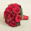 Resting on a softly textured beige cloth that resembles delicate linen, this compact bouquet of deep red roses immediately draws the eye with its intensity and poise. Dozens of velvety blooms are arranged in a perfectly rounded dome, each head nestled closely to the next, creating a sense of abundance and unity. The rose petals show a spectrum of deep scarlet, crimson, and wine-red tones, some with slightly ruffled edges that hint at movement, as though a gentle Oxford breeze from the banks of the Thames had just passed over them. Threaded between the roses are glossy red hypericum berries, small and spherical, catching the light and adding lively points of contrast against the softness of the flowers. Encircling the entire arrangement, dark green leaves are folded and wrapped with deliberate care, forming a tailored collar that holds the bouquet together and enhances its sculpted silhouette. The stems are hidden beneath a band of bright red satin ribbon, tightly wound to form a smooth, elegant handle stretching toward the right of the image. The elevated viewpoint lets you see the symmetry, the careful spacing of berries, and the exacting craftsmanship you might expect from a local Oxford florist preparing bridal flowers for a ceremony in Summertown or a romantic celebration near Radcliffe Square. The combination of rich reds, glossy greens, and the calming neutral background evokes warmth, intimacy, and timeless romance, making the bouquet ideal for weddings, anniversaries, or same-day romantic deliveries across Oxford's historic streets.