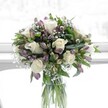 A graceful, full bouquet of creamy white roses, lavender and muted purple alstroemeria, and airy white baby's breath stands in a clear cylindrical glass vase, illuminated by soft natural daylight that feels like it's drifting through an Oxford townhouse window. The white roses are fully open yet still perfectly formed, each velvety petal curling outward to create a plush, romantic centre that immediately catches the eye. Around them, clusters of alstroemeria buds and half-open flowers in gentle shades of lilac and smoky purple, veined with deeper tones, weave an intricate pattern of colour and texture that feels both modern and timeless. Tiny clouds of gypsophila float between the larger blooms, lending a feathery, cloud-like softness that lightens the whole design. Lush green foliage, from broad, deep-toned leaves to slimmer, more delicate stems, frames the flowers and gives the arrangement its rounded, carefully balanced shape. Through the crystal-clear vase, you can see the neatly trimmed green stems, immersed in fresh water, aligned in a way that reveals the florist's precise, practiced hand. The overall palette is calm and serene, reminiscent of a quiet walk along the River Thames in Oxford at first light, with pale colours gently emerging from the morning mist. This bouquet feels designed for moments that ask for tenderness and sincerity-perhaps a thoughtful sympathy gesture delivered to a home off Iffley Road, a gentle congratulations for a new beginning in Jericho, or a simple way to bring peace and soft fragrance into a sitting room near the Covered Market-an arrangement that speaks in a whisper of comfort, admiration, and quiet celebration.