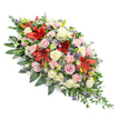 A dignified, double-ended casket spray is shown against a bright white studio background, its gentle diagonal line guiding the viewer's gaze from one end to the other, as though following the path of a final farewell through Oxford's tree-lined streets. The arrangement is densely packed yet thoughtfully balanced, combining traditional blooms with more contemporary touches. Deep red Oriental-style lilies stand out as commanding focal points, their large, star-shaped petals radiating warmth and quiet strength. They are surrounded by a generous selection of roses in cream, white, and soft pink, some fully open and others in half-bud, their smooth, layered petals creating a sense of tenderness and devotion. Pale pink spray carnations appear throughout, their ruffled heads adding softness and volume, while small white and daisy-like flowers nestle between larger blooms, brightening the overall palette. Cool tones are introduced through small spikes of blue-purple thistles and airy clouds of lavender-blue filler flowers, likely statice or veronica, lending texture and depth much like the varied architecture around Carfax and the High Street. The flowers sit on a lush cushion of deep green foliage composed of broad, polished leaves and trailing, feathery greenery that extends slightly beyond the blossoms, creating a natural frame and a sense of movement. The lighting in the image is even and gentle, allowing the reds, pinks, whites, and purples to appear vivid but not harsh. This long, elegant spray feels designed to be viewed from all sides, perfect for resting atop a coffin or adorning a memorial, expressing heartfelt condolences and the enduring love of family and friends gathering to say goodbye.