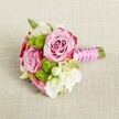 A small, hand-tied bouquet rests horizontally on a softly textured, light tan fabric that looks like natural linen, the neutral background allowing every flower to stand out clearly. At the centre, two fully opened pink roses and a third partly hidden bloom form the heart of the design, their petals moving from gentle blush in the middle to deeper, almost magenta tones at the ruffled outer edges, like a delicate ombré. Their spiralled centres appear velvety and intricate, suggesting that just moments ago they were still unfurling. Nestled closely around the roses are rounded tufts of vivid lime green button chrysanthemums, each one a neat, spherical cluster of tiny petals that add playful texture and a modern twist to the romantic pinks. Between these focal blooms, creamy white and soft ivory filler flowers - reminiscent of lisianthus, freesia, or wax flower - cascade lightly through the bouquet, their small, star-shaped blossoms and tiny yellow centres introducing a sense of movement and freshness. Slender hints of deep green foliage peek through, grounding the colours and giving a garden-fresh feel, as if gathered from a border in an Oxford townhouse courtyard. The stems are neatly gathered and trimmed, then wrapped in a smooth fuchsia pink satin ribbon that catches the light with a gentle sheen. A slim strand of white faux pearls is carefully wound around the handle in a diagonal criss-cross, evoking bridal jewellery and adding an air of quiet luxury. Soft, even lighting bathes the bouquet, with no harsh shadows, making it easy to imagine this compact posy in the hands of a bridesmaid walking through an Oxford college chapel or used as an intimate table arrangement at a small celebration near the Covered Market, radiating romance, elegance, and a sense of poised anticipation.