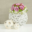 On a pristine white tabletop that catches and mirrors the light, a striking bridal-style centerpiece takes pride of place, the kind you could imagine gracing a boutique Oxford wedding venue near the Sheldonian or along St Giles. The arrangement forms a gentle, rounded dome of flowers, sitting securely within a spherical vase woven from slender, white-painted twigs. This vessel has an open, airy pattern, each twig looping and twisting so that the structure feels both sculptural and feather-light, adding a modern rustic note. Above it, pastel pink roses in full bloom create the main visual melody. Their petals curl generously outwards, some tipped with deeper blush tones, suggesting softness and warmth. Nestled among them are vivid lime-green button chrysanthemums, their tight, textured discs breaking up the smoothness of the rose petals and introducing a fresh, lively green. At the base, fine white flowers with tiny yellow centers gather like a cloud, partially hiding the rim of the vase, and giving the impression that the bouquet gently floats above the woven branches. To the left, two decorative orbs constructed from pale wooden ribbons echo the circular theme, their natural colour grounding the composition. The backdrop is a softly blurred, linen-like wall in warm taupe, which provides quiet depth without competing for attention. The entire arrangement speaks of thoughtful craftsmanship and gentle celebration, perfect for a wedding breakfast table, an engagement dinner in an Oxford town house, or a refined reception overlooking the college gardens, where fresh flowers are chosen to complement rather than dominate the setting.