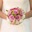 Held close against the intricate lace of a bridal gown, this bouquet captures the tender calm just before an Oxford wedding begins. The eye is first drawn to the lush lavender-pink roses, their colour hovering between mauve and dusty blush, with velvety petals that spiral inward in flawless symmetry. Tucked among them are bright lime-green button chrysanthemums, their round, cushion-like blooms offering a lively contrast that lifts the whole arrangement into something distinctly contemporary. Along the outer edge, dainty white flowers and pointed buds form a soft, ruffled border, echoing the fine lace and tulle often seen in bridal boutiques along the High Street. The bouquet is round and hand-tied, sitting snugly yet naturally in the bride's hands, the stems neatly wrapped so that only the fresh green tips peek out. In your mind, you can almost breathe in the fragrance-a gentle mix of rose petals and cool greenery, reminiscent of early walks through Christ Church Meadow when the air is still and hopeful. The light in the image is soft and flattering, like late morning sunshine filtering between Oxford's honey-stone buildings, catching the subtle sheen on each petal. Every detail speaks of skilled craftsmanship and thoughtful design, the colours chosen to stay elegant in photographs from the ceremony at a city church to celebrations in a college hall or riverside marquee along the Thames. It is a bouquet that feels reassuring and refined, a quiet statement of joy and new beginnings.