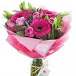This hand-tied bouquet reads like a joyful spring promise: several large fuchsia gerbera daisies with deep, almost black centers form the visual anchor, their broad, velvety petals radiating intense colour. Around them nest delicate pink roses in varying stages-some open, their layers of soft petals unfurling like satin spirals; others kept as refined buds that suggest a slow, fragrant reveal. Sprays of deep purple-blue statice weave subtle texture between the focal blooms, their tiny clustered florets offering cool contrast against the hot pinks. Lush green foliage-broad, glossy leaves and slender, verdant stems-frames the flowers and adds depth, the greens grounding the composition with an earthy clarity. The bouquet is swathed in crinkled, sheer pink wrapping that graduates from an almost translucent blush halo at the top to a richer, saturated pink lower down, catching the light with a mix of glossy gleam and soft translucence. The tied stems rest in a clear cylindrical glass vase, visible through the water, and a piece of rustic twine loops round the vase neck where a small white rectangular tag hangs, embossed with a simple pink-and-green rose motif that hints at a local florist's care. Light on the scene is gentle and true, like a bright morning in Oxford's Covered Market, bringing out the sheen on the petals and the tactile depth of the cellophane. As an arrangement this suits birthdays, Mother's Day, or a fresh congratulations-an exuberant, carefully balanced gift that smells faintly sweet and green, and reads as cheerful, generous, and unmistakably handcrafted.
