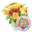 A bright, comforting 'Get Well Soon' gift captured against a clean white backdrop: a lush glass-vased bouquet on the left and a shiny round Mylar balloon to the right, both arranged to lift the spirits of someone recovering. The bouquet is generous and loose, with several large, soft orange-to-deep-yellow roses opening at the center; their velvety petals curl outward and suggest a faint, citrus-tinged fragrance. Surrounding the roses are sunny yellow gerbera daisies whose broad faces and dark centres give an immediate sense of cheer, while rich burgundy-to-maroon carnations add ruffled texture and visual weight. Small clusters of white alstroemeria (Peruvian lilies) peek between the bolder blooms, lending delicate points of contrast and a slight sweetness, and abundant glossy green foliage frames the flowers so the composition reads as fresh and verdant. Stems and clear water are visible through the simple glass vase, emphasizing floral freshness and care in handling. To the lower right, partially overlapping the stems, a metallic silver Mylar balloon gleams; its reflective surface carries a playful central design of cartoon balloons in blue, purple, green, pink and yellow with stripes, polka dots and checks, and bold yellow-and-blue lettering declaring the message Get Well Soon. Tiny starburst motifs scatter around the balloon's white rim and a small Kaleidoscope logo sits unobtrusively at the bottom edge. Bathed in soft daylight, the ensemble radiates warmth, compassion and a hopeful uplift-perfect for sending to a friend or neighbour convalescing in and around Oxford.