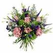 A romantic, garden-style bouquet fills the frame, its colours and textures heightened by a simple white background that makes each bloom appear freshly gathered from an Oxfordshire meadow. At the centre, you can almost feel the softness of pastel pink roses, their petals unfurling in delicate spirals from deeper rosy hearts to paler, creamy edges. Around them, generous carnations in dusty rose and mauve create waves of ruffled texture, adding a sense of movement and gentle opulence, as if the flowers are caught mid-sigh. Piercing through this softness, tall spires of Veronica in a deep violet-blue rise like elegant brushstrokes, lending height and a slightly untamed energy reminiscent of the borders lining the paths around Christ Church Meadow. Tucked between these vertical elements are velvety purple lisianthus blooms, their bell-shaped petals folded and layered with quiet sophistication. Threaded throughout the bouquet are airy sprays of tiny, star-like aster blooms in lavender and periwinkle, creating a soft, misty veil that lightens and opens the design. A rich tapestry of greenery encircles the flowers: lime-green textural filler adds bright, lively flashes of colour, while muted, silvery eucalyptus leaves and deeper green foliage keep the palette balanced and calm. The hand-tied stems are visible at the base, neatly bundled and evenly cut, ready to slip into a favourite vase in a Jericho townhouse or a cosy terrace near Cowley Road. The arrangement exudes a mood of thoughtful celebration and quiet affection, ideal for same-day delivery in Oxford when words alone do not feel enough.