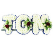 In this photograph, the name TOM is beautifully spelled out in three-dimensional floral letters, the kind of bespoke tribute often chosen for a funeral service or wake in and around Oxford. Each capital letter is densely covered with pure white chrysanthemums, their small blooms packed side by side to create a velvety, cushion-like surface that looks soft to the touch and symbolises peace and remembrance. Tracing the outline of every letter is a royal blue satin ribbon, its clean edge and gentle shine giving the design a tailored, formal finish and helping the name stand out clearly when viewed in a church, cemetery, or crematorium. Carefully arranged at key points on the T, O, and M are compact floral sprays composed of deep red roses, miniature white or cream spray roses, and graceful, white freesia-style flowers. These are nestled into clusters of lush green foliage, including fern and other textured leaves, which add depth, colour contrast, and a natural, garden-like feel. The letters sit aligned on a slim dark support frame, suggesting they are ready for delivery by a local Oxford florist to a service perhaps near Headington, Botley, or one of the city's peaceful churchyards. The overall impression is one of quiet dignity and personal affection: white blooms for purity and farewell, blue ribbon for serenity and respect, and red roses for love that endures beyond loss. This floral arrangement honours Tom's name with grace, providing a comforting focal point for family and friends during a time of grief.
