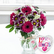 A warm, joyful still life unfolds in soft daylight, as a vibrant bouquet and a heart-shaped balloon share a spotless, pale tabletop, evoking the feeling of a surprise delivery arriving at a home near Oxford's High Street. The arrangement is full and rounded, gathered in a sturdy, clear glass vase that reveals the cleanly cut stems and the clear water that keeps them fresh. At the heart of the bouquet, several striking gerbera daisies in a rich magenta hue open like cheerful faces, their dark central discs giving each bloom depth and drama. Surrounding them is a lively ring of smaller chrysanthemums, their petals starting as deep, velvety purple at the base before gently lightening to bright white at the tips, each flower crowned with a glowing yellow centre. Nestled between these statement blooms are elegant creamy white roses, some still tightly furled, others beginning to unfurl their softly scented petals, adding a timeless, romantic touch. Fine sprays of white baby's breath weave through the flowers, like delicate lace, softening edges and creating a sense of airiness, while fresh green leaves peek out throughout, adding a natural, garden-like frame. Around the neck of the vase, a length of rustic, natural twine is tied, holding a small white tag printed with a pink rose motif and friendly green script that hints at lovingly chosen, hand-picked flowers from a caring local florist. Slightly to the right, leaning into the display, a shiny foil balloon shaped like a heart glints with silver edging. Its white centre is covered with a mosaic of colourful letters in pinks, purples, and blues spelling out Love n' Hugs 24-7, echoing the bouquet's playful, affectionate spirit. The softly blurred, bright background suggests a window and a calm, airy room, making this scene perfect for imagining a thoughtful, same-day floral surprise sent to cheer someone in Headington, Cowley, or the city centre.