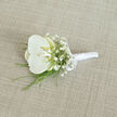 A refined white orchid boutonnière rests at a gentle angle on a pale, linen-coloured fabric, its elegance revealed in soft, consistent light that picks up every subtle texture. At the heart of the design is a single orchid bloom, pristine and sculptural, its velvety petals fanning out in rounded, overlapping layers of pure white. In the centre, a hint of warm cream and muted yellow glows softly, drawing the eye toward the flower's throat without disturbing the overall simplicity. Encircling this focal blossom, clusters of white baby's breath appear like tiny stars against a clouded sky, their numerous miniature blooms adding height, airiness, and an almost weightless texture. Fine strands of bright green foliage extend beneath and behind the flowers, slender and graceful, giving a sense of movement and natural freshness reminiscent of riverside greenery along the Thames path near Oxford's Folly Bridge. The stems are carefully aligned and tightly wrapped in sleek white satin ribbon, creating a polished handle that would sit smoothly against a tailored jacket. A petite satin bow, crafted from the same ribbon, is secured near the top of the binding, its neat loops and short tails conveying meticulous craftsmanship. The background fabric, a subtly woven light beige or tan, offers a neutral, earthy stage for the boutonnière, with its visible weave echoing the feel of high-quality suiting. This image captures the quiet luxury of a wedding day detail-perfect for a groom, best man, or honoured guest at an Oxford ceremony-embodying calm sophistication and the gentle romance of fresh, hand-prepared flowers.
