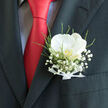 A refined wedding boutonnière rests inside the breast pocket of a charcoal suit, its design centred on a single white orchid that seems to glow softly against the dark fabric. The orchid's petals are broad, creamy, and smooth, with gentle curves that catch the light like fine bone china, and at its heart a hint of pale gold suggests warmth and tenderness. Nestled around this focal bloom is a delicate halo of baby's breath, each tiny white flower forming a cloud-like border that softens the composition, reminiscent of the light Oxford mist that can drift along the High Street on early mornings. Slender stems of greenery and wisps of fern weave through the gypsophila, adding movement and a subtle, natural rhythm that feels as if it could have been gathered from a secluded corner of Port Meadow. A slim white satin ribbon is tied precisely at the base of the arrangement, its sheen echoing the orchid's quiet elegance and hinting at the careful hands that crafted it. The boutonnière contrasts beautifully with the clean lines of the suit and the vivid splash of a red tie, whose silk sheen introduces a note of celebration and bold personality. This floral accent feels as though it belongs at a joyful Oxford wedding, perhaps in a historic college hall or a chic venue near Jericho, where tradition meets contemporary style. It speaks of love expressed in understated detail, of promises made beneath ancient spires, and of a day where every petal, texture, and colour has been thoughtfully chosen to honour the moment.