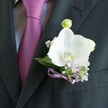 Pinned neatly above a crisp pocket on a dark tailored jacket, this white orchid boutonniere feels like the finishing note to a carefully composed occasion in Oxford, perhaps a ceremony tucked behind old stone walls or a reception near St Giles. The orchid itself is luminous, its smooth, ivory-tinged petals unfolding in graceful arcs like a calm breath before walking down the aisle. At the centre, a touch of buttery cream adds depth, as though the flower holds a secret warmth. Encircling it, cool, silvery-green leaves and soft lilac accents create a halo of texture, their muted tones chosen to enhance, not overshadow, the central bloom. Tiny lavender sprigs and white buds peek through, recalling the delicate wildflowers that edge paths along the Isis in early spring. A narrow violet ribbon binds the stems with clean, even turns, the satin catching the light in slender flashes that mirror the purple-pink tie beneath. The overall composition is compact yet layered, thoughtfully proportioned so it sits close to the fabric without feeling heavy or fussy. Light glances across the arrangement, picking out the curve of each petal and the quiet shimmer of the ribbon, suggesting freshness and the immediacy of the moment. It feels like a piece created by a florist who understands both tradition and modern style-someone who knows how to make a groom, best man, or proud father feel subtly distinguished as they step into a day of vows, toasts, and new memories in the heart of Oxford.