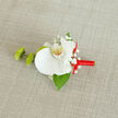 On a neutral, light beige fabric backdrop that suggests woven linen or soft burlap, a handcrafted boutonnière is laid out as if waiting to be pinned to a tuxedo before an Oxford town hall ceremony. At its heart, a single white orchid unfurls pristine, velvety petals, the surface silky and smooth, catching the light in gentle, satiny reflections. The center of the orchid glows with subtle yellow and orange speckles, a small but striking detail that draws the eye inward. Around this focal bloom float tiny clusters of white baby's breath, each miniature flower contributing a cloud-like texture that softens the composition and adds a note of quiet romance. On the left, two bright green buds with a textured, slightly spiky surface-reminiscent of green button chrysanthemums-press close to the orchid, offering a fresh, modern contrast in form and colour. Beneath the arrangement, a single deep green leaf, glossy yet understated, acts as a natural base, anchoring the floral elements like foliage glimpsed in an Oxford garden after rain. The stem is expertly wrapped in a smooth, vibrant red satin ribbon, spiralling neatly before ending in a small bow that arcs to the right, its radiant hue symbolising warmth, love, and celebration. The boutonnière feels both timeless and contemporary, ideal for weddings at one of the historic colleges, proms, or formal dinners along Banbury Road. Every element appears thoughtfully positioned, speaking to the practiced hands of a local florist who understands how a modest lapel flower can quietly express commitment, style, and personal story.