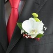 A close-up image shows a wedding boutonniere carefully pinned to the lapel of a dark charcoal suit jacket, the fine wool fabric revealing a soft, woven texture under natural, diffused light. At the heart of the design is a single, pristine white Phalaenopsis orchid, its broad, velvety petals opening like wings and framing a delicate yellow-and-white lip that draws the eye. Just above the orchid sit two petite, lime-green button chrysanthemum blooms, their rounded forms and feathery petals adding a bright, contemporary touch of colour. Around them, clusters of tiny white baby's breath create a light, cloud-like halo, softening the lines and lending an airy, romantic feel. A few rich, glossy green leaves cradle the flowers, giving structure and a sense of freshness, as if they've just been prepared in a florist's workshop off the High Street in central Oxford. The base of the boutonniere is wrapped in a slim, deep red satin ribbon, its sheen subtly echoing the bold, solid red satin tie that hangs to the left against a pale grey and white checked dress shirt. The contrast between the dark suit, the bright tie, and the tranquil white orchid captures the quiet formality of a wedding morning, perhaps at a ceremony near Oxford's historic colleges. Every detail of the image speaks of calm anticipation and thoughtful craftsmanship, the kind of finishing touch a groom might choose for a refined, modern celebration while walking through Broad Street or heading to a reception along the river.