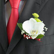 Pinned to the lapel of a classic black suit, this orchid boutonnière is captured in close detail, showing the care and craftsmanship behind a seemingly simple design. At its centre is a pristine white orchid, the petals thick and smooth like polished ceramic, opening in a relaxed, elegant curve. A soft touch of buttery yellow glows at the throat of the flower, adding warmth and depth. Behind the orchid, a pair of lime-green button chrysanthemums stand slightly taller, their round, tightly packed petals giving a pleasing, cushion-like texture that contrasts with the orchid's sleek surface. Wisps of delicate baby's breath weave around the main blooms, each tiny white flower no larger than a pinhead, creating a misty, lace-like border. A narrow ribbon in deep, romantic red wraps the stems at the base, tying the composition together and subtly mirroring the rich red tie that runs down the front of the suit. The dark jacket fabric provides a matte backdrop, making the clean whites and greens feel bright and fresh, like a walk through University Parks on a crisp spring morning in Oxford. The foliage, deep green and tidy, supports the flowers without stealing attention, reinforcing the boutonnière's sense of structure and calm. The whole arrangement feels tailored and personal, ideal for a groom or groomsman stepping into a ceremony at a chapel off High Street or a reception near Jericho. Crafted with an eye for proportion and texture, it is the kind of discreet floral detail that elevates formalwear without overpowering it.