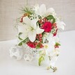 A sophisticated floral centerpiece, perfect for an Oxford wedding table, is displayed against a soft, light beige backdrop that enhances its calm, romantic character. The arrangement sits in a round, open-weave white basket, whose airy wickerwork pattern adds a contemporary yet handcrafted feel, as if sourced from a local Oxford artisan market off Cornmarket Street. Rising gracefully from this base, large white oriental lilies dominate the design, their broad, velvety petals fully open and gently curved, showing off tall stamens tipped with warm pollen. Interwoven with the lilies are rich, velvety red roses, tightly layered and intensely colored, introducing a sense of passion and timeless romance. Fine sprigs of white baby's breath drift between the larger flowers like soft clouds, while light, feathery greenery and bright green leaves create gentle movement and a fresh, garden-like feel, akin to a stroll through University Parks on a crisp day. Small clusters of glossy red hypericum berries nestle among the blooms, catching the light and adding a festive, seasonal dimension ideal for Christmas weddings or winter anniversary dinners. Extending from the right side, a delicate cascade of white phalaenopsis orchids arches outward, their rounded petals creamy and luminous, with subtle yellow and blush tones at the centers, and a few buds still closed, promising future blossoms. On the left side of the arrangement, three white woven spheres are placed on the white tabletop, echoing the texture of the main container and visually balancing the scene. The overall impression is of refined elegance and intimate celebration, a design that would feel perfectly at home in an Oxford college dining hall, a boutique hotel near the Bodleian, or a romantic dinner setting in Jericho.